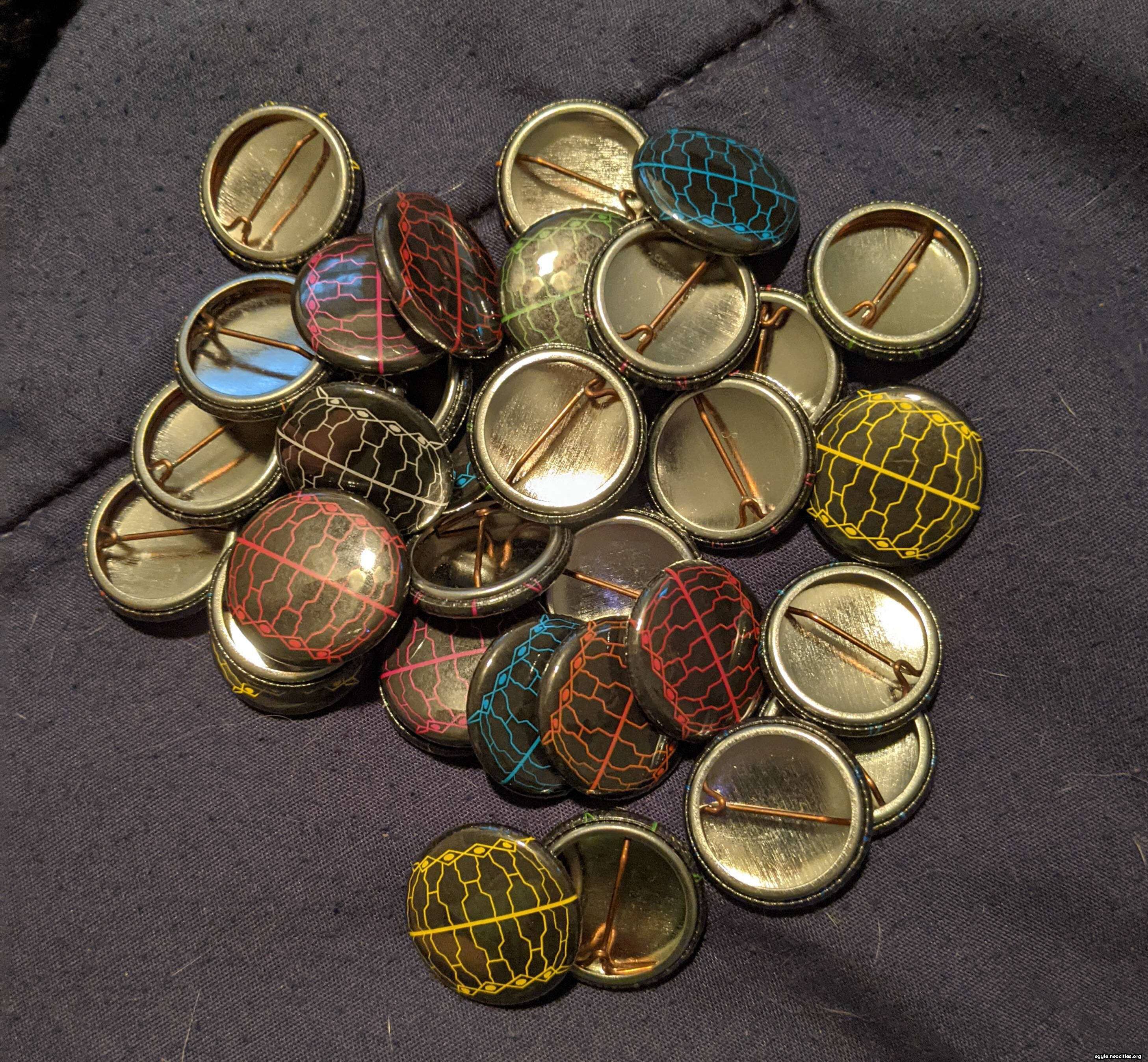 A pile of one inch circular metal pinback buttons. The featured design is an Allagan Companion Node minion in different colors.