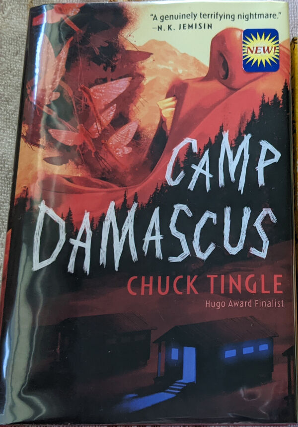 Camp Damascus book cover, written by Chuck Tingle, hugo award finalist. The cover art features two camp cabins, one with blue lights coming from the door and windows. Behind them is a dark treeline of evergreens, and behind the trees is the bottom half of a large human face, the mouth open as if screaming, with several mayflies flying out. A quote at the top from N. K. Jemisin reads: A genuinely terrifying nightmare. In the upper left corner is a library sticker that is blue wiht a yellow sunburst that says New.