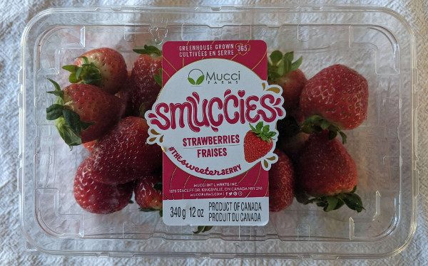 A closed plastic clamshell container of strawberries. The Logo sticker reads: Greenhouse grown, Mucci Farms Smuccies Strawberries. the sweeter berry. Product of Canada. The label also has the same phrases in french.