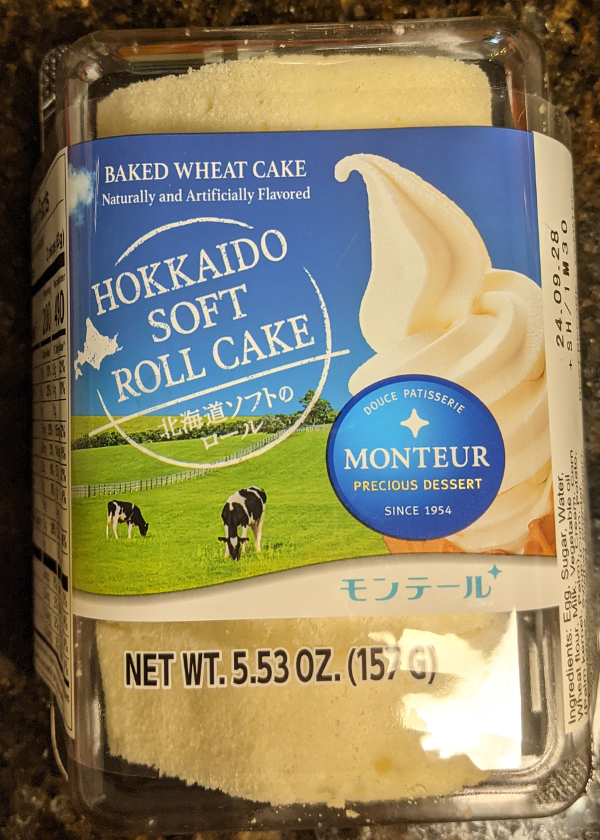 Monteur Hokkaido Soft Roll cake in packaging. The label shows a cone of soft serve vanilla ice cream in the foreground. In the background is a lush green grassland with two black and white cows grazing. The text on the label reads Baked Wheat Cake, Naturally and artificially flavored. Hokkaido Soft Role Cake. There is a logo which reads Dolce Patisserie Monteur. Precious Dessert Since 1954.