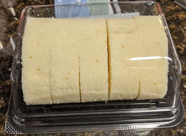 The roll cake in its packaging, a black tray covered by a transparent plastic dome, seen from the side