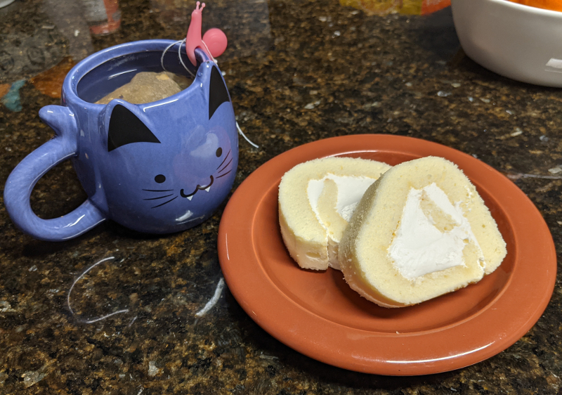 A slice of roll cake on a small brown plate. To the left is a purple mug that looks like a smiling cat with vampire fangs. A tea bag is floating at the top. On the rim of the mug is a small silicone snail meant to hold the tea bag string so it does not fall into the cup.