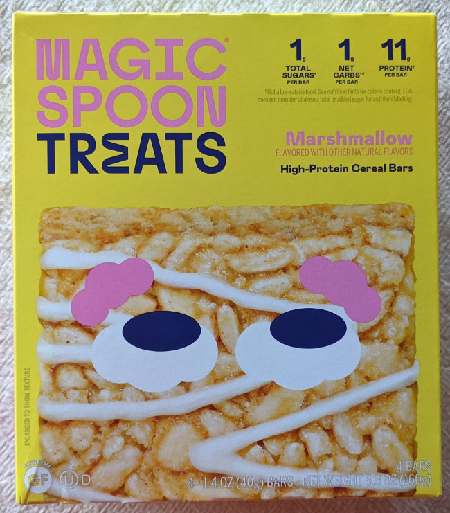 A box of Magic Spoon Treats in marshmallow flavor. The box is yellow with a cereal treat at the bottom that has eyes with pink eyebrows. The eyes are looking towards the informational text in the corner. Text on the box reads: Magic Spoon Treats. 1g total sugar, 1g net carbs, 11g protien per bar. Marshmallow Flavored with other natural flavors. High Protien cereal bars. 4 1.4 OZ (40g) bars. Gluten Free.