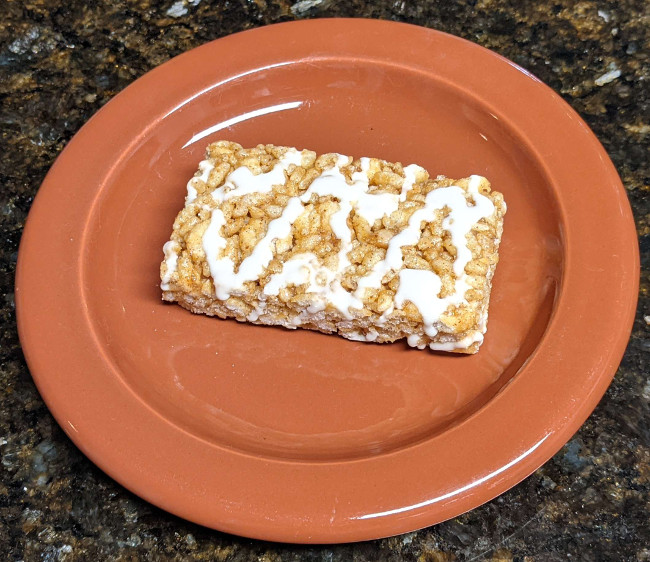 A marshmallow treats bar sitting on a small brown plate. It looks similar to a rice crispy treat bar, and has a drizzle of white icing on top.