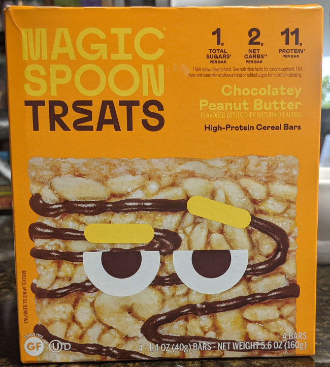A box of Magic Spoon Treats in chocolatey peanut butter flavor. The box is orange with a cereal treat at the bottom that has bored looking eyes with yellow eyebrows. Text on the box reads: Magic Spoon Treats. 1g total sugar, 2g net carbs, 11g protien per bar. Chocolatey Peanut Butter Flavored with other natural flavors. High Protien cereal bars. 4 1.4 OZ (40g) bars. Gluten Free.
