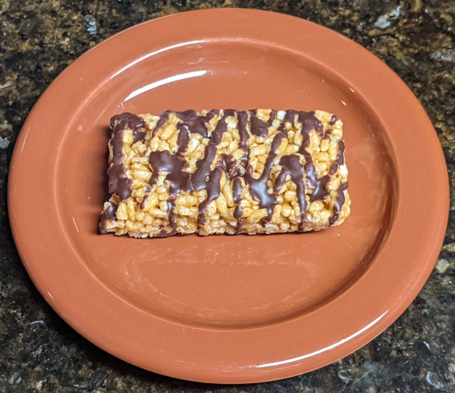 A chocolate peanut butter treats bar sitting on a small brown plate. It looks similar to a rice crispy treat bar, and has a drizzle of chocolate on top.